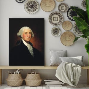 May include: A framed portrait of George Washington in a dark suit with a white cravat. The portrait is hanging on a white wall with a collection of woven wall decor.