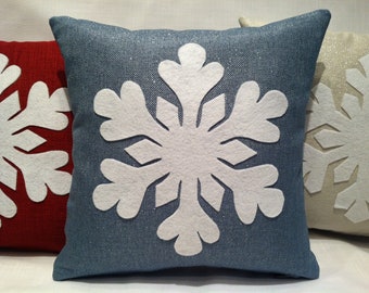 May include: Three decorative throw pillows with snowflake designs. The pillows are made of a textured fabric and have a white snowflake appliqué on a red, blue, and cream background.