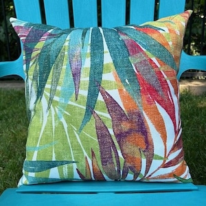 May include: A colorful throw pillow with a tropical leaf pattern in shades of green, orange, red, and purple. The pillow is on a blue chair.