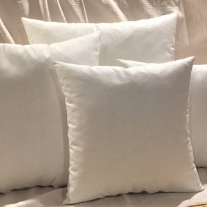 May include: Five white square pillows arranged in a staggered formation on a light beige fabric surface. The pillows are all the same size and shape, and they are all made of a soft, plush material.