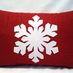 May include: Red pillow with a white snowflake design. The snowflake is made of felt and is centered on the pillow.