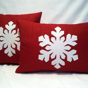 May include: Two red throw pillows with white felt snowflake designs. The pillows are made of a textured fabric.