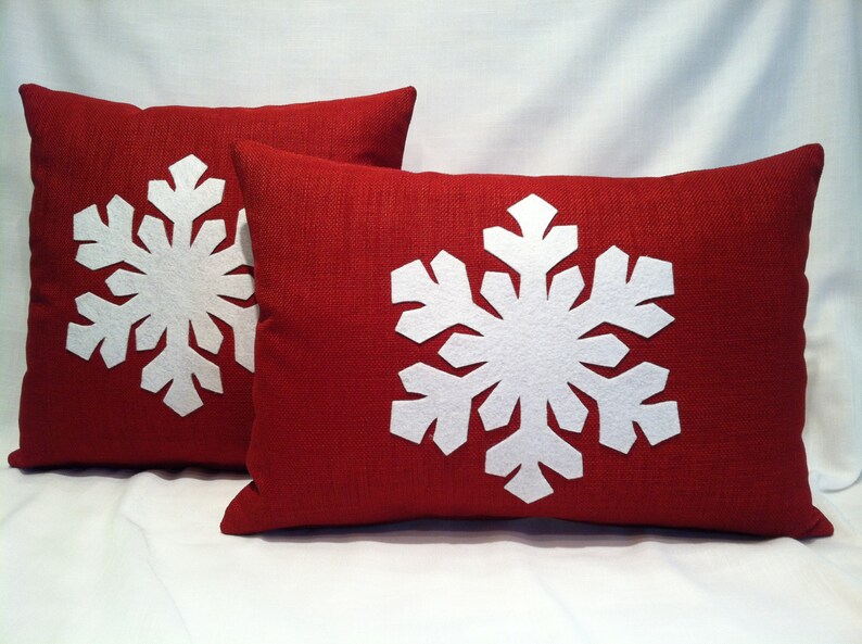 May include: Two red throw pillows with white felt snowflake designs. The pillows are made of a textured fabric.