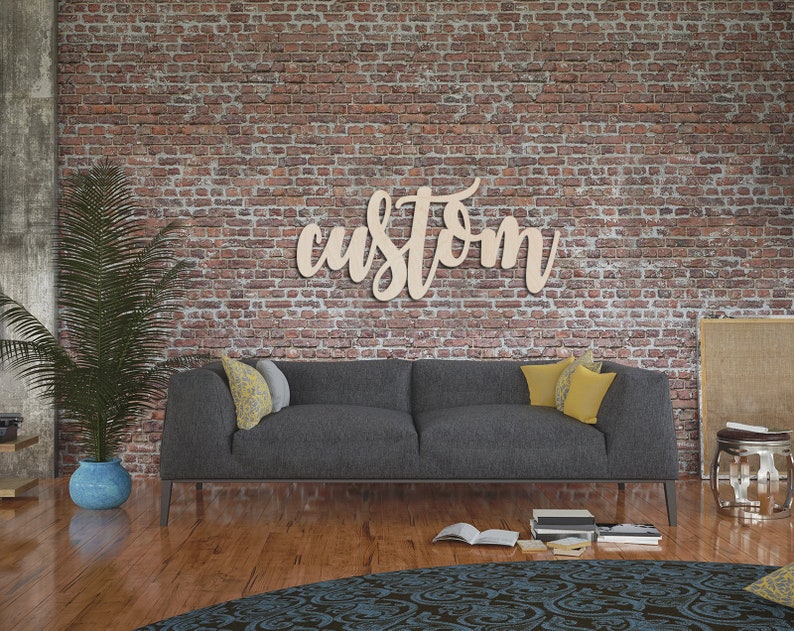 May include: A wooden sign that reads "custom" in a cursive font is mounted on a brick wall. A gray sofa with yellow and gray pillows sits below the sign. A potted plant and a patterned rug are in the foreground.