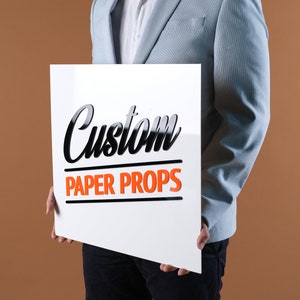 May include: A person holding a white sign with black text that says "Custom Paper Props".