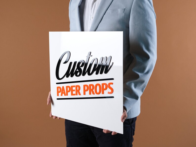 May include: A person holding a white sign with black text that says "Custom Paper Props".