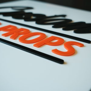 May include: A white sign with black lettering that reads "Custom Props". The lettering is in a bold, stylized font. The words "Custom Props" are in orange raised letters.
