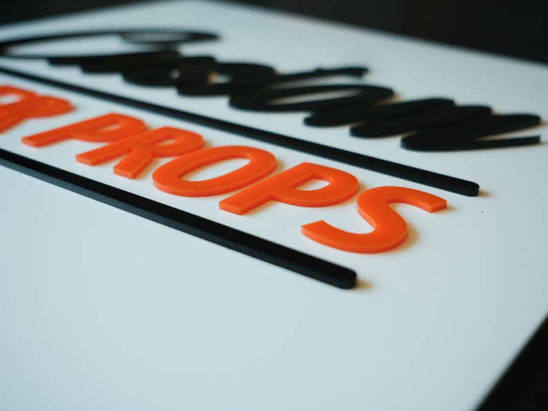 May include: A white sign with black lettering that reads "Custom Props". The lettering is in a bold, stylized font. The words "Custom Props" are in orange raised letters.