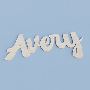 May include: Wooden cutout of the name "Avery" in a cursive font. The letters are made of light-colored wood and are set against a solid light blue background. The name is a decorative element.