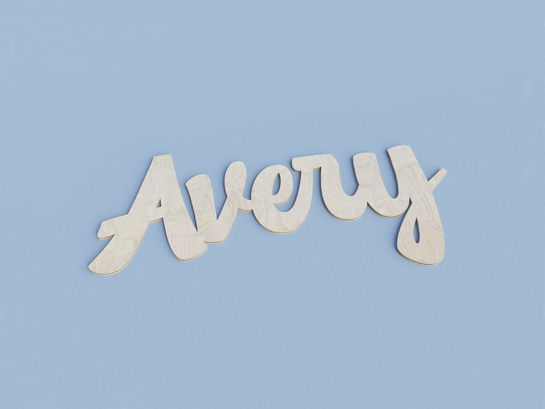 May include: Wooden cutout of the name "Avery" in a cursive font. The letters are made of light-colored wood and are set against a solid light blue background. The name is a decorative element.