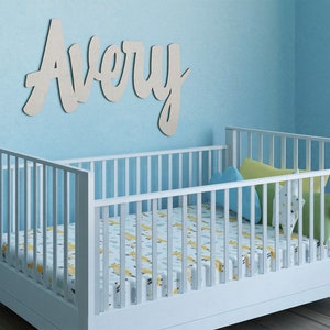 May include: A white crib with a patterned sheet and pillows is in a room with a light blue wall. The name "Avery" is displayed in cursive lettering above the crib. The crib is made of wood.