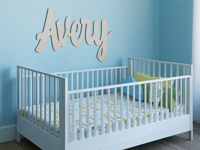 May include: A white crib with a patterned sheet and pillows is in a room with a light blue wall. The name "Avery" is displayed in cursive lettering above the crib. The crib is made of wood.