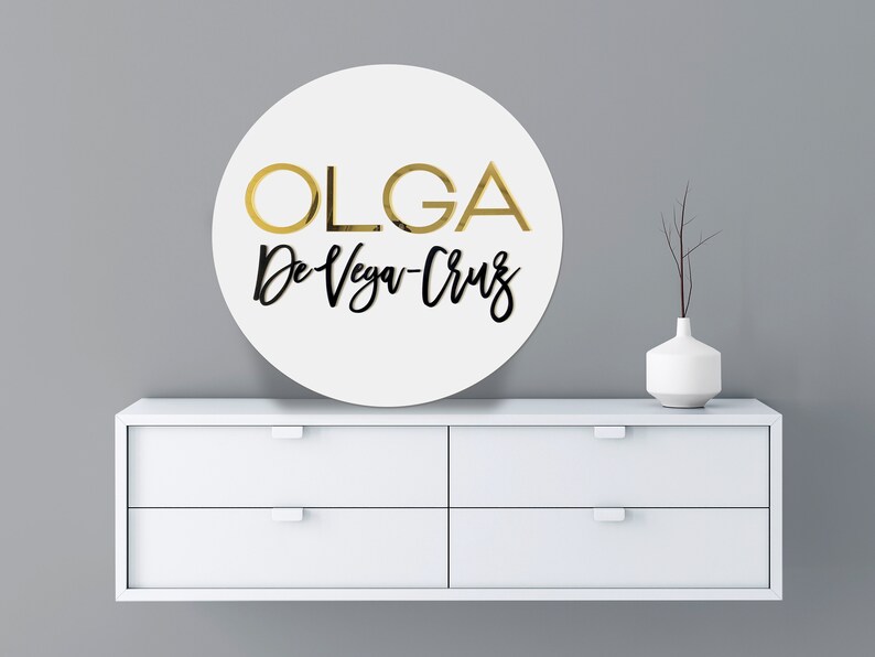 May include: A white round sign with the text "OLGA De Vega-Cruz" in gold and black lettering.