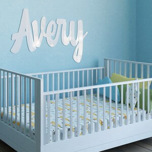 May include: White wooden crib with a white and yellow patterned sheet. The crib is in a room with light blue walls and a white wooden name sign that says "Avery" on the wall.
