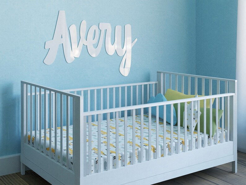 May include: White wooden crib with a white and yellow patterned sheet. The crib is in a room with light blue walls and a white wooden name sign that says "Avery" on the wall.