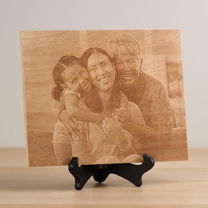 May include: A wooden engraved photo plaque with a portrait of three generations of a family. The plaque features a grandmother, mother, and daughter smiling and hugging each other. The plaque is on a small black stand.