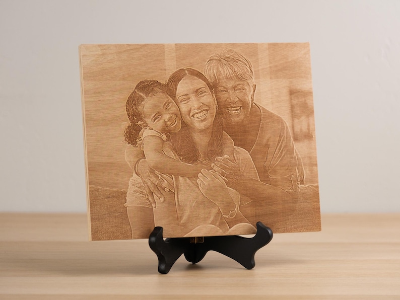 May include: A wooden engraved photo plaque with a portrait of three generations of a family. The plaque features a grandmother, mother, and daughter smiling and hugging each other. The plaque is on a small black stand.