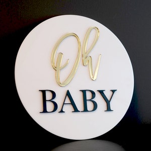 May include: A white circular sign with the words "Oh Baby" in gold and black lettering. The "Oh" is in a cursive script font and the "Baby" is in a block font.