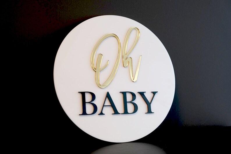 May include: A white circular sign with the words "Oh Baby" in gold and black lettering. The "Oh" is in a cursive script font and the "Baby" is in a block font.
