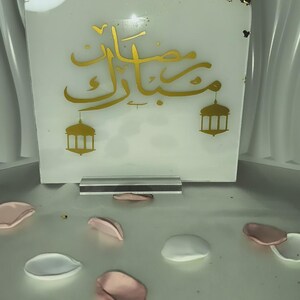 May include: A white rectangular decorative piece with gold Arabic calligraphy and two lantern illustrations. Gold leaf accents the top edge. The piece is propped up on a clear stand, with scattered pink and white petals in the foreground.
