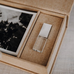Puede incluir: Una caja de lino beige con una foto en blanco y negro de un ramo de flores y una unidad USB de cristal.