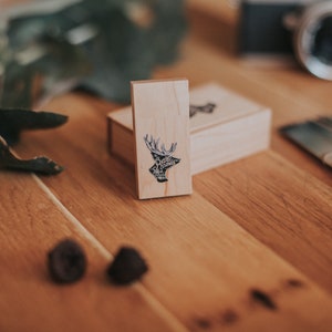 Puede incluir: Una caja de madera con un diseño de cabeza de ciervo en la parte delantera. La caja está hecha de madera clara y tiene un acabado natural. La cabeza de ciervo es negra y blanca y tiene un diseño simple.