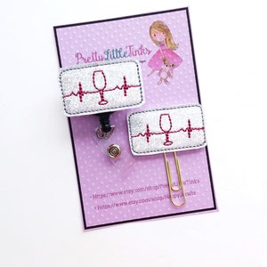 May include: Two white glitter badge reels with a red wine glass and a red heartbeat design. The badge reels are on a pink background with white polka dots. The text "Pretty Little Tinks" is at the top of the image. The text "https://www.etsy.com/shop/PrettyLittleTinks" and "https://www.etsy.com/shop/Happy2Crafts" is at the bottom of the image.
