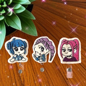 May include: Three embroidered badge reels featuring cartoon characters with blue, purple, and pink hair. Each badge reel has a different character design, with a white border and a retractable cord for attaching to clothing or accessories.