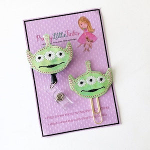 May include: A green alien character with three eyes, embroidered on a fabric badge, with a gold clip. The badge is on a purple background with white polka dots. The text "Pretty Little Tinks" is visible in the top right corner of the image. The text "https://www.etsy.com/shop/PrettyLittleTinks" and "https://www.etsy.com/shop/Happy2Crafts" is visible at the bottom of the image.