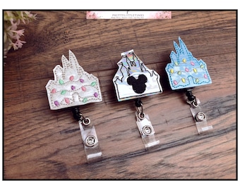 May include: Three Disney castle themed retractable badge holders. The first is white with silver glitter and light blue and pink lights. The second is iridescent with a black Mickey Mouse silhouette. The third is light blue with yellow lights.