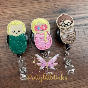 May include: Three embroidered badge reels with character designs. One has a green outfit, one a pink outfit, and the third has a brown outfit with glasses. A clear badge holder with a butterfly design is in the center, with the text "prettylittletinks".