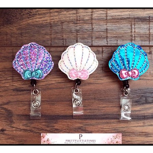 May include: Three sparkly seashell-shaped badge holders with pink bows. The shells are purple, white, and blue.
