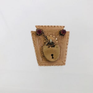 May include: A gold-toned locket pendant with a heart-shaped lock and keyhole. The locket is decorated with a chain and two red gemstones.