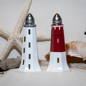 May include: Two ceramic lighthouse salt and pepper shakers. The shaker on the left is white with black accents. The shaker on the right is white with red and black accents.