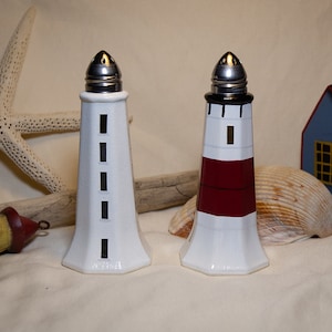 May include: Two ceramic lighthouse salt and pepper shakers with silver tops. One is white with black rectangular windows, the other is white with a red stripe. The background includes a starfish, shell, and small wooden house.