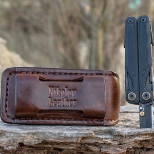 Puede incluir: Una funda de cinturón de cuero marrón con el texto en relieve "Nikolov Leather" junto a una multiherramienta negra. La funda es rectangular con una trabilla para el cinturón. La multiherramienta es negra con una hoja parcialmente expuesta.