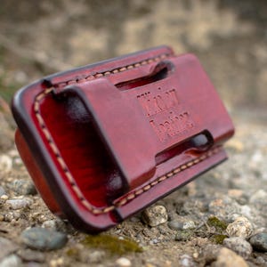 Pode incluir: Uma carteira de couro bordô com costuras visíveis e o texto em relevo "Iron Leather". A carteira é retangular com bordas arredondadas e parece estar sobre pequenas pedras.