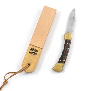 Könnte beinhalten: Ein Holzstrop mit der Aufschrift "Nikolov Leather" und einem Lederriemen neben einem Klappmesser. Das Messer hat eine Klinge aus rostfreiem Stahl, Messingbacken und einen dunklen Holzgriff.