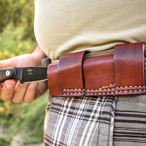 Könnte beinhalten: Ein braunes Leder-Messer-Etui mit einem genähten Design, das an einem beigefarbenen Gürtel getragen wird. Das Etui hat eine Klappe, die das Messer sichert.