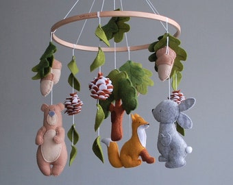 May include: A hanging nursery mobile featuring a circular wooden hoop with white strings suspending felt figures of a brown bear, an orange fox, and a gray rabbit, interspersed with green felt leaves, brown acorns, and small pinecones.