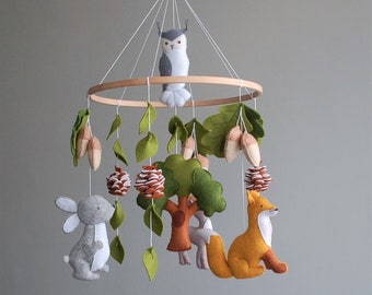 May include: A hanging nursery mobile featuring a circular wooden frame with felt figures including a grey owl, a grey rabbit, an orange fox, a brown tree, green leaves, tan acorns, and white and brown pinecones suspended by white strings.