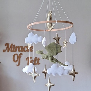 Pode incluir: Um m&oacute;bile suspenso com um drag&atilde;o central de feltro verde-claro rodeado por nuvens de feltro branco e estrelas e uma lua crescente em dourado met&aacute;lico, suspensos por uma estrutura circular de madeira com fios brancos. Texto Miracle Of Felt.