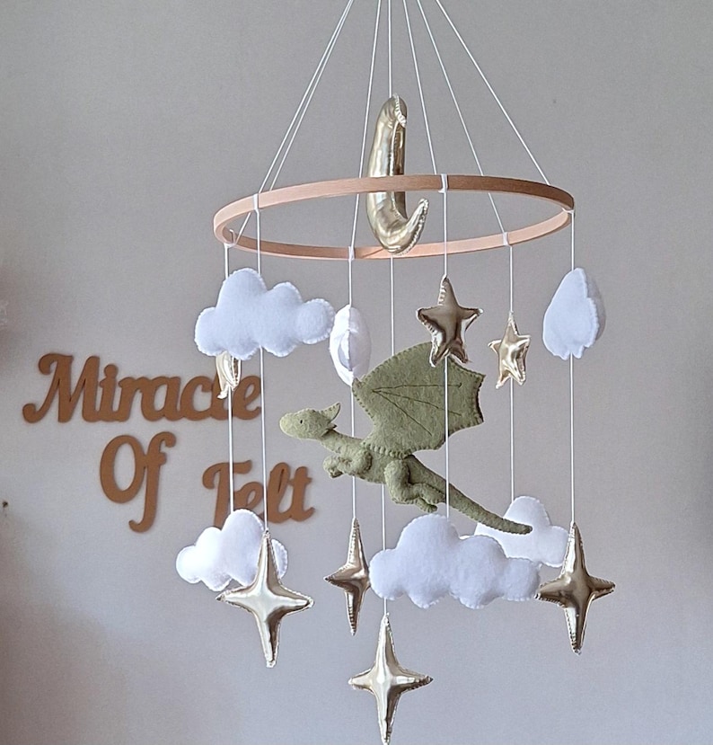 Pode incluir: Um m&oacute;bile suspenso com um drag&atilde;o central de feltro verde-claro rodeado por nuvens de feltro branco e estrelas e uma lua crescente em dourado met&aacute;lico, suspensos por uma estrutura circular de madeira com fios brancos. Texto Miracle Of Felt.