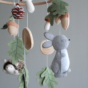 May include: A nursery mobile featuring felt woodland elements including a gray rabbit, a hedgehog, brown acorns, a pinecone, a mushroom, and green oak leaves, all suspended from a wooden hoop by white strings, interspersed with small wooden discs.