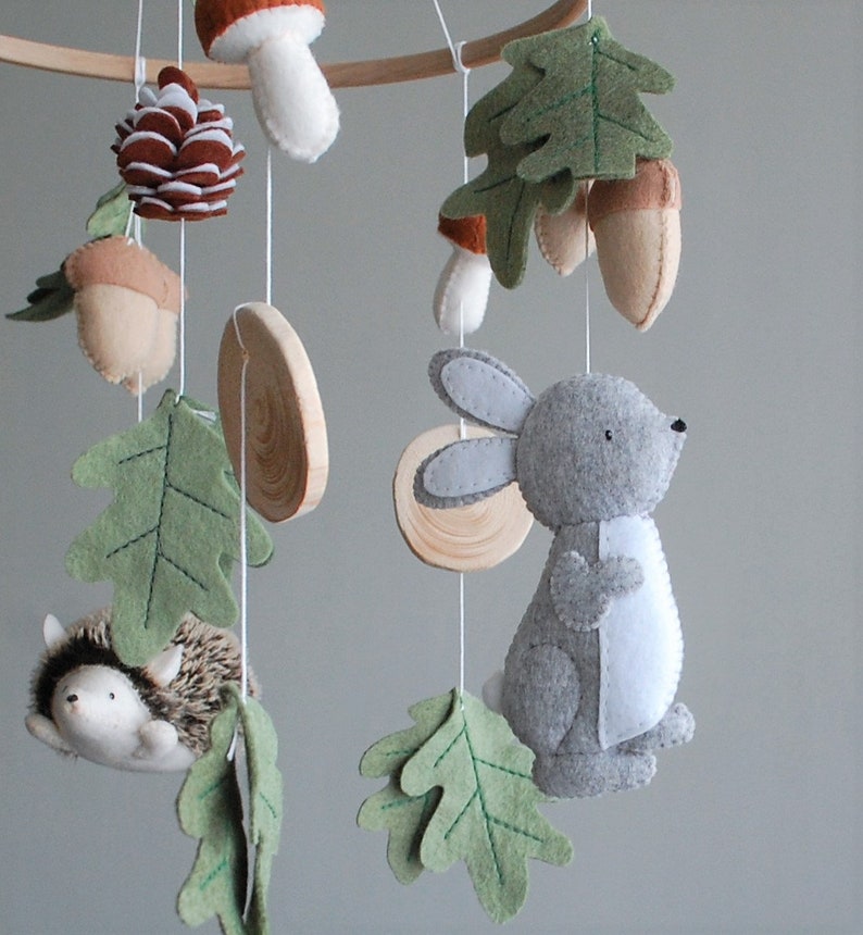 May include: A nursery mobile featuring felt woodland elements including a gray rabbit, a hedgehog, brown acorns, a pinecone, a mushroom, and green oak leaves, all suspended from a wooden hoop by white strings, interspersed with small wooden discs.