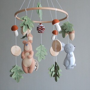 May include: A hanging nursery mobile featuring a circular wooden frame with felt figures including a tan bear, a gray rabbit, orange and white mushrooms, green oak leaves, acorns, and a pinecone, all suspended by white strings from the frame.