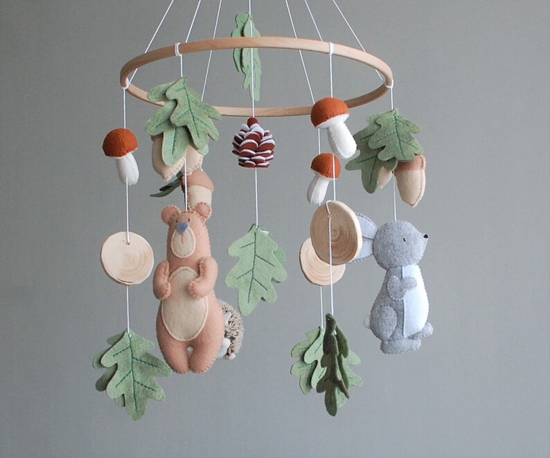 May include: A hanging nursery mobile featuring a circular wooden frame with felt figures including a tan bear, a gray rabbit, orange and white mushrooms, green oak leaves, acorns, and a pinecone, all suspended by white strings from the frame.