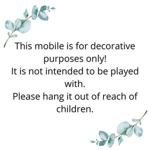 Puede incluir: Un letrero rectangular blanco con ramas de eucalipto en acuarela en las esquinas superior izquierda e inferior derecha. El texto negro dice: This mobile is for decorative purposes only! It is not intended to be played with. Please hang it out of reach of children.