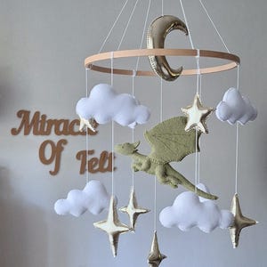 Pode incluir: Um m&oacute;bile suspenso com um drag&atilde;o central de feltro verde-claro, rodeado por nuvens de feltro branco e estrelas e uma lua crescente em dourado met&aacute;lico, tudo suspenso numa estrutura circular de madeira por fios brancos finos.