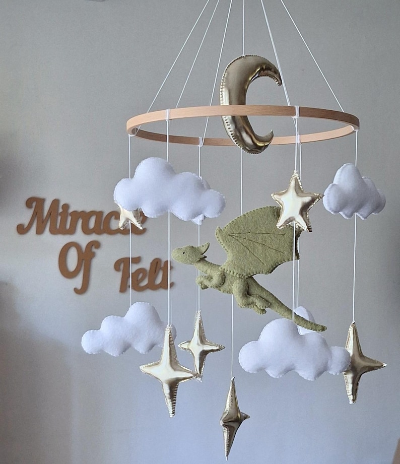 Pode incluir: Um m&oacute;bile suspenso com um drag&atilde;o central de feltro verde-claro, rodeado por nuvens de feltro branco e estrelas e uma lua crescente em dourado met&aacute;lico, tudo suspenso numa estrutura circular de madeira por fios brancos finos.